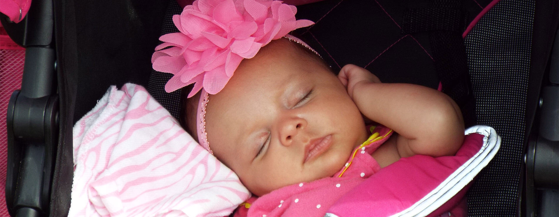 A sleeping baby in her car seat. She is snuggling a pink blanket and is wearing a pink hair band.